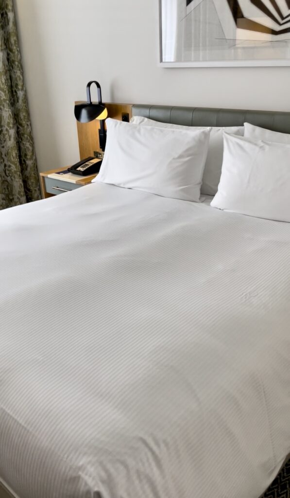 Neatly made hotel bed with luxurious white linens and modern side tables, set against patterned curtains and contemporary wall art. Shows a luxury price point option at a dog friendly Dublin hotel.