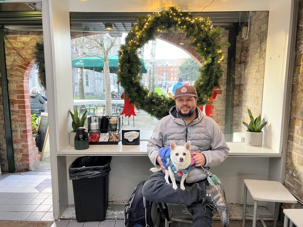 Man sitting inside a cozy cafe in front of a large holiday wreath, holding a small white dog on his lap.