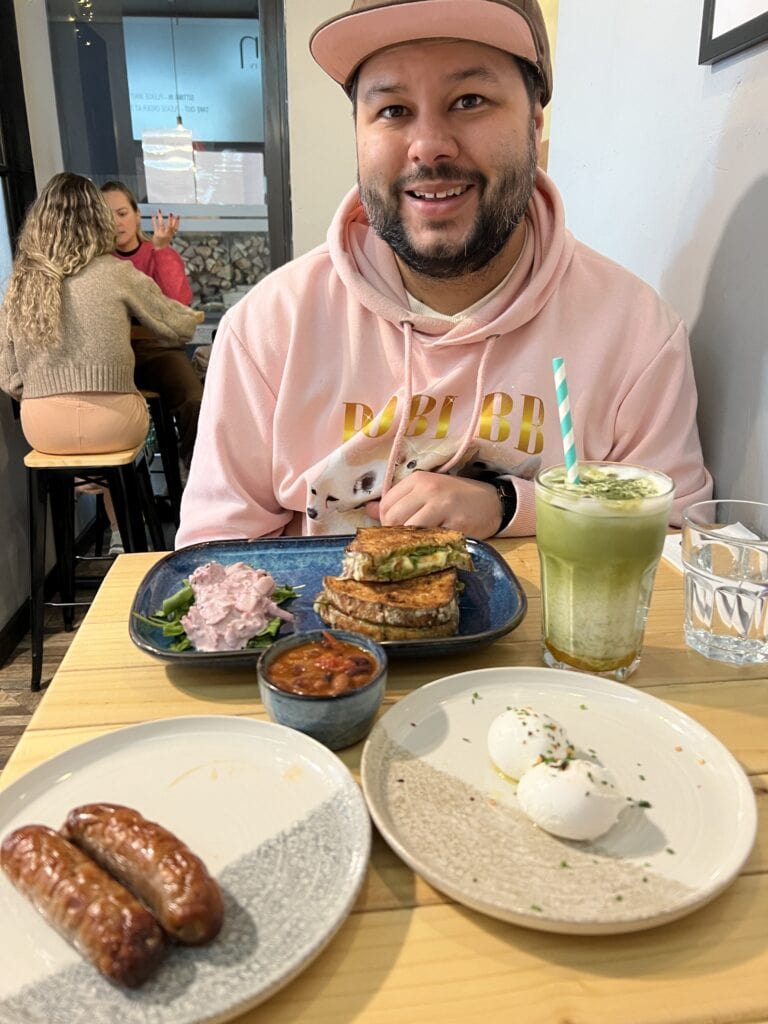 A smiling man in a pink hoodie with dog illustrations sits at a wooden table with a brunch spread including grilled focaccia, poached eggs, sausages, beans, and a mango matcha at a dog-friendly café in Dublin.