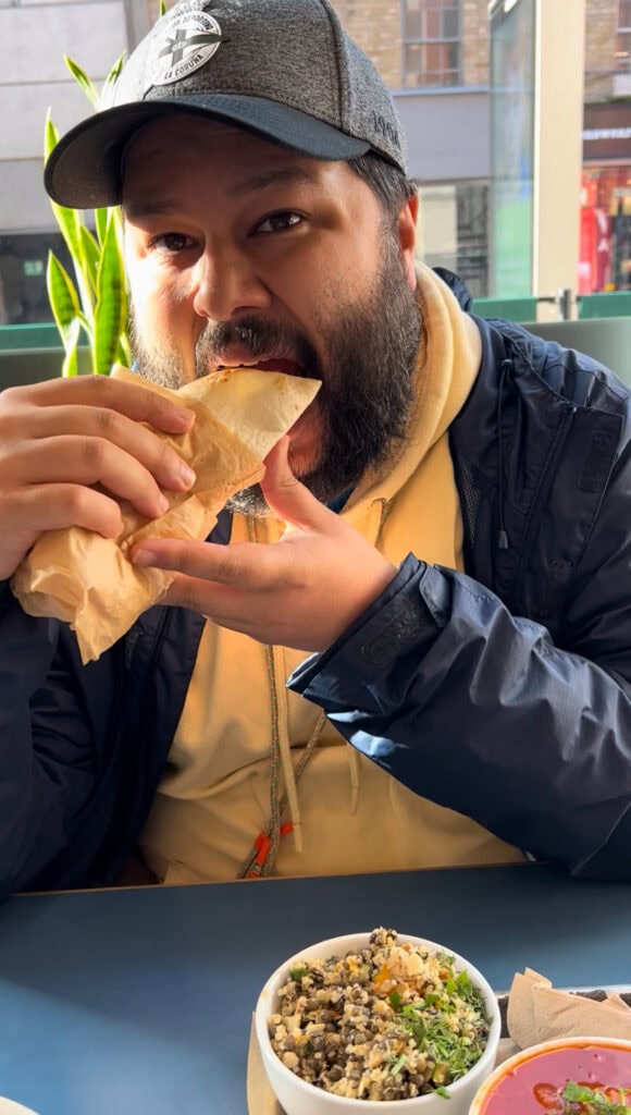 Man bites intro a wrap at a sunny table inside a Dublin brunch spot, with bowls of salad and soup in front of him.