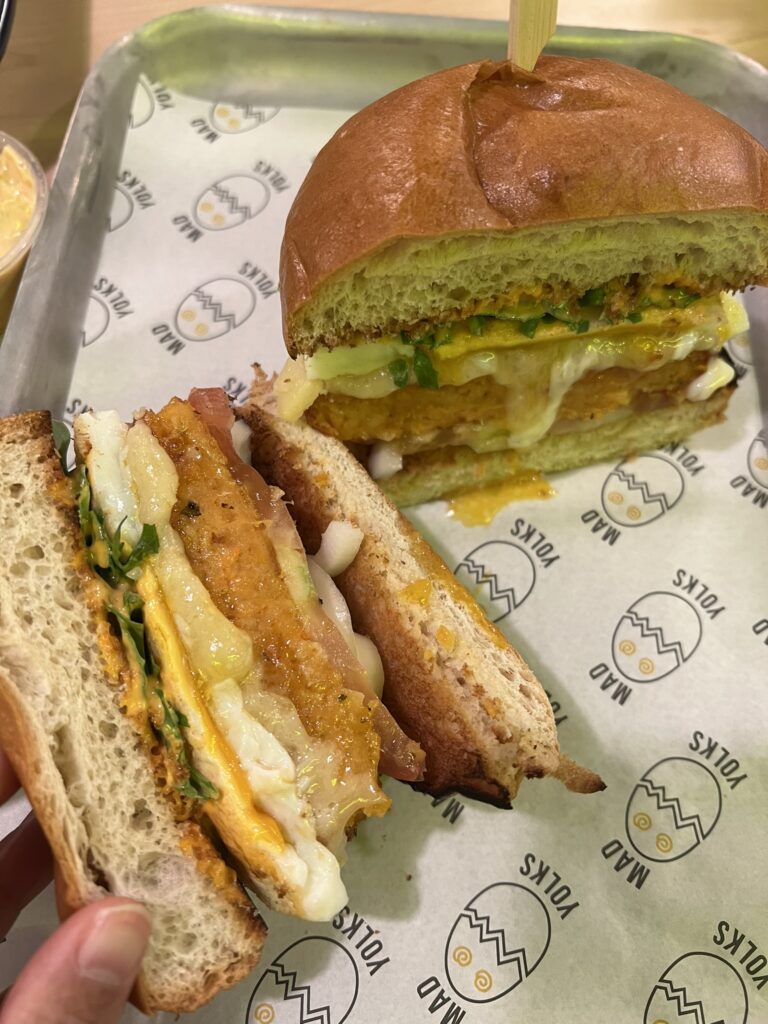 Cross-section of a gourmet breakfast sandwich with egg, cheese, and hash brown on a toasted bun, served on Mad Yolks branded paper.