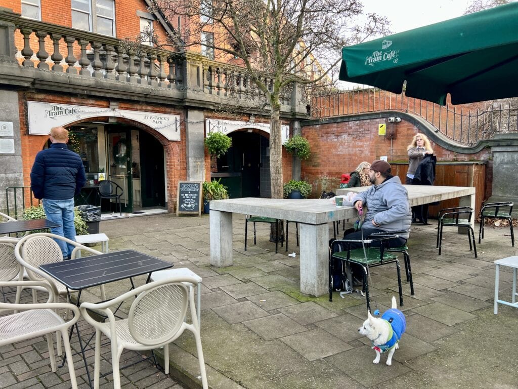 Outdoor seating area of The Tram Café in Dublin with a man seated and a small white dog in a blue coat standing nearby.