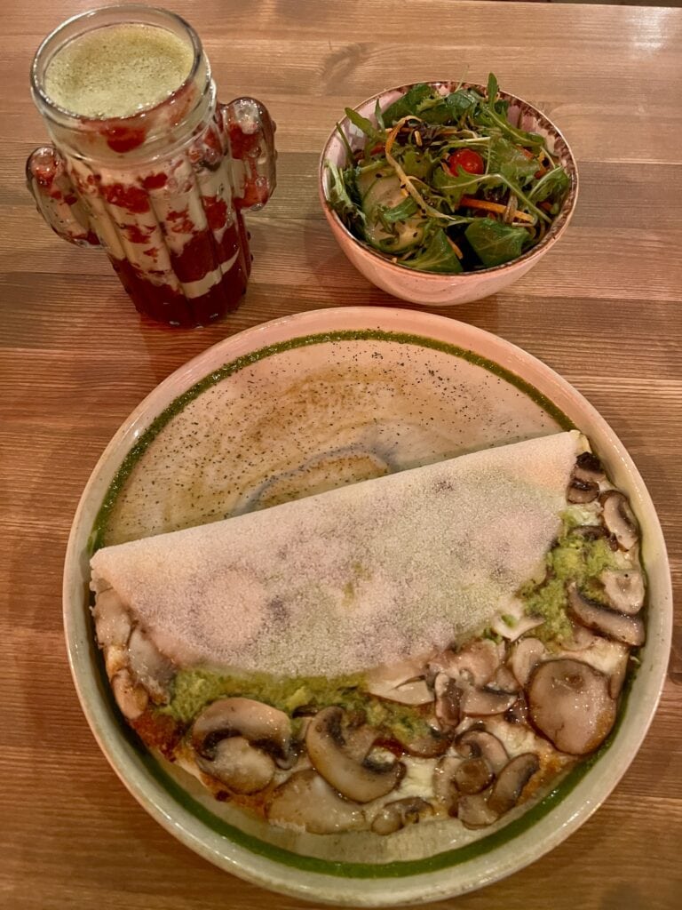 Overhead shot of a tapioca wrap filled with cheese, mushrooms and avocado, served with a fresh green salad and an iced strawberry matcha drink in a trendy cactus-shaped glass.