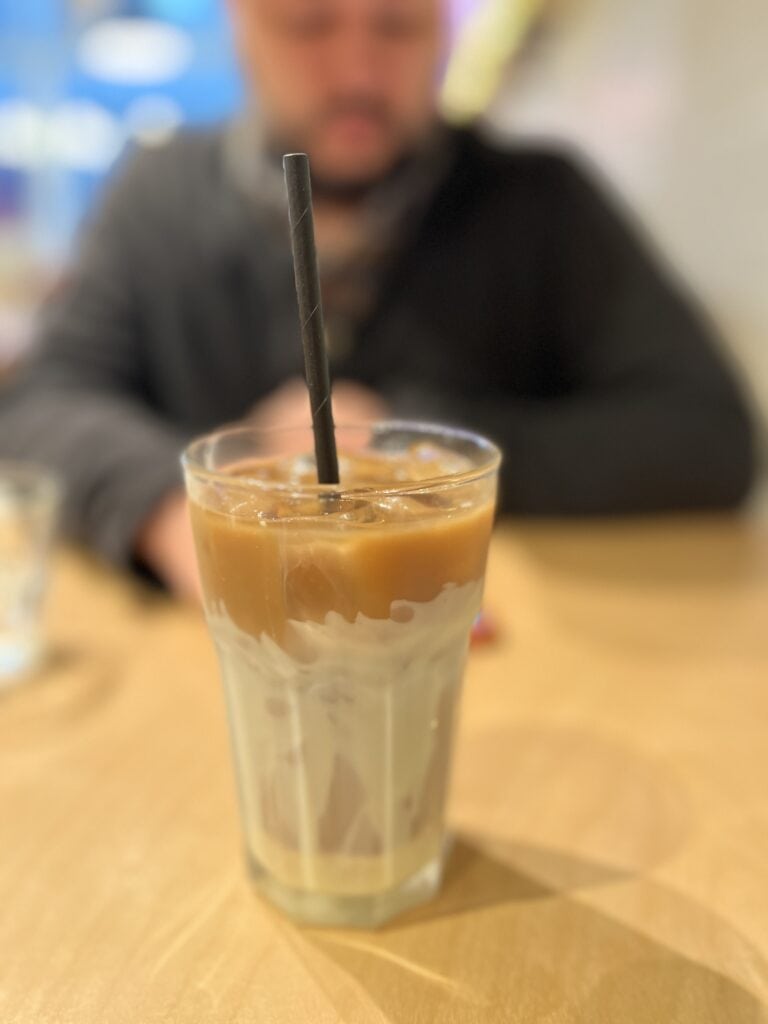 A close-up of a Vietnamese iced coffee latte (ca phe sua da) in a clear glass with a black straw, set on a wooden table. The background is softly blurred, showing a seated man out of focus.