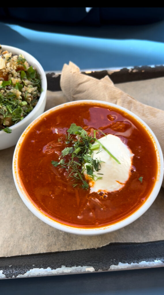 Close-up of a vibrant bowl of beetroot potato borscht topped with fresh herbs and a dollop of sour cream, served alongside a quinoa and lentil salad.