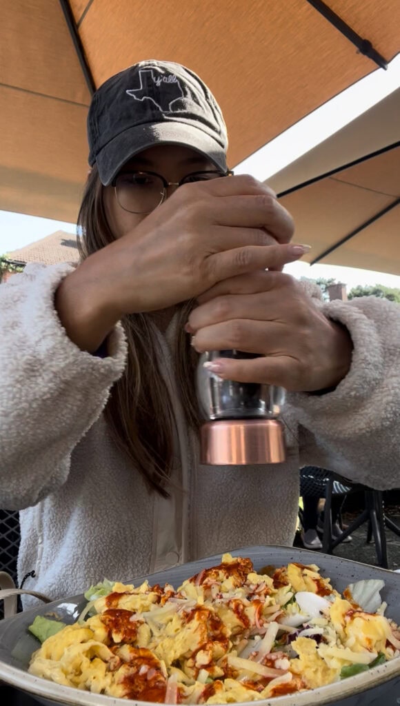 Woman in a fleece jacket and baseball cap grinding fresh pepper over a bowl of scrambled eggs and salad during an outdoor brunch at a dog-friendly café in Dublin.