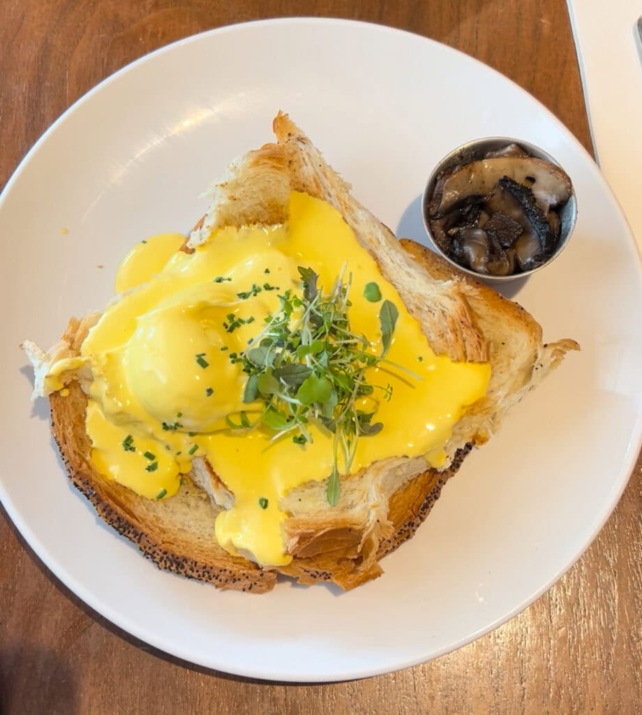 A rich eggs Benedict-style brunch dish with runny hollandaise sauce over toast, topped with microgreens and served with a side of sautéed mushrooms.