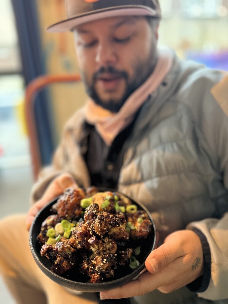 A man in a baseball cap holds a bowl of fried teriyaki glazed cauliflower bites topped with sesame seeds and scallions at the Japanese cafe Sando Paradiso.