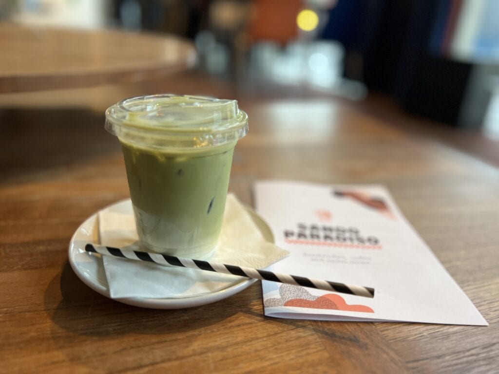 A cup of iced matcha latte on a small plate with a striped straw, next to a Sando Paradiso menu on a wooden table.