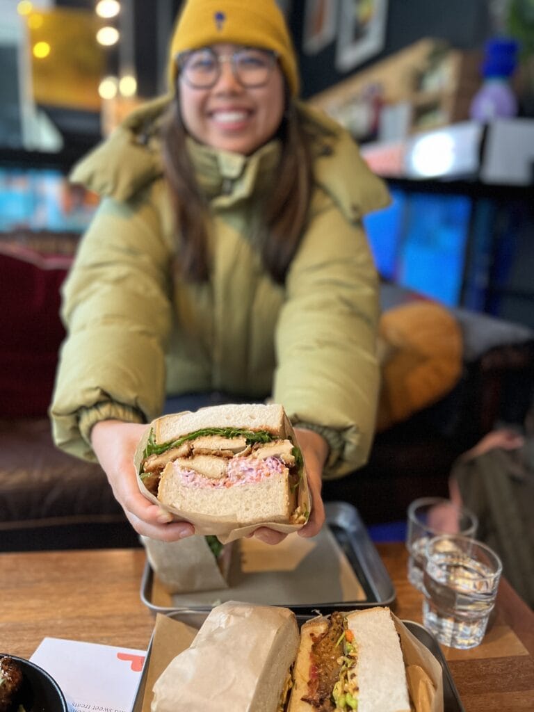 A smiling woman in a yellow beanie and green jacket holds up a thick Japanese-style katsu chicken sando. The sandwich is made with panko fried chicken, shredded slaw, kewpie mayo and tonkatsu sauce on three inch thick bread at the japanese cafe Sando Paradiso in Dublin.